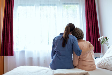 Female Home Nurse Hugging Elderly Woman on Bed. Back View of Female Nurse With Her Arm Around Elderly Patient Shoulder. - obrazy, fototapety, plakaty