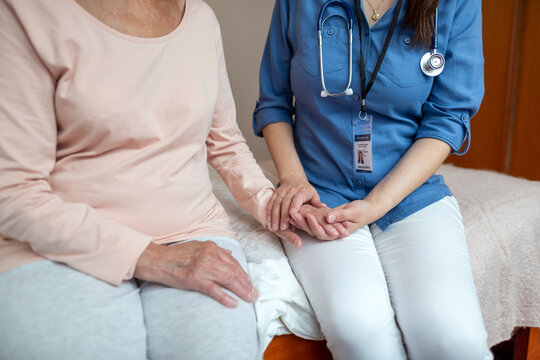 Close up of Home Caregiver Holding Hands of Elderly Woman Patient. Female Nurse Sitting On Hospital Bed with Patient Caressing Her Hands.