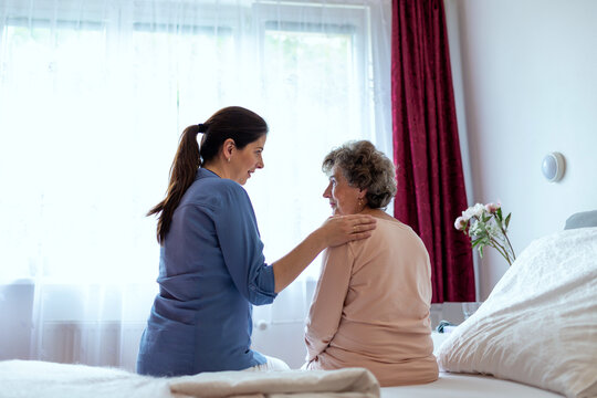 Home Caregiver Comforting Senior Woman Patient Sitting in Her Bedroom. Nurse Putting Her Hand Over Elderly Patient Shoulder in Hospital Room.