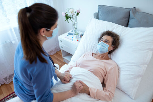 Elderly Woman Wearing Protection Face Mask Looking at Nurse Leaning Over. Top View of Home Caregiver With Face Mask Holding Hands of Female Senior Patient Lying in Bed at Nursing Home.