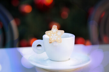 Violet bokeh on white porcelain coffee cup with saucer and wooden decorative star with snowflakes. Blurred background with chairs and Christmas tree with red balls. New year postcard. Congratulations.