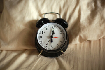Top view of classic black alarm clock showing six o'clock with a bed in the background, waking up in the morning concept