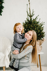 Mother and little child having fun and playing together at home. Portrait loving family close up. Cheerful mom hugging cute baby daughter girl near Christmas tree. Merry Christmas and Happy Holidays.