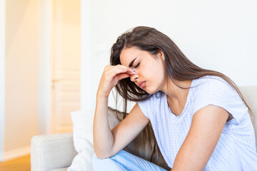 Portrait of an attractive woman sitting on a sofa at home with a headache, feeling pain and with an expression of being unwell. Upset depressed woman sitting on couch feeling strong headache migraine.