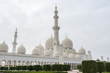 Fototapeta na wymiar Minaret and domes of white Grand Mosque against white cloudy sky, also called Sheikh Zayed Grand Mosque, inspired by Persian, Mughal and Moorish mosque architecture