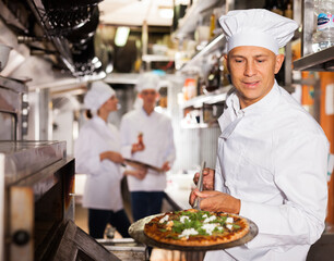 Qualified chef standing near professional oven in modern restaurant kitchen, holding in his hands shovel with cooked pizza..