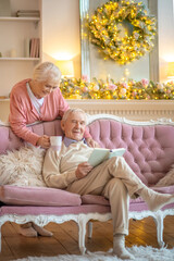 Elderly man sitting on a sofa with a book and his wife giving him a cup of tea