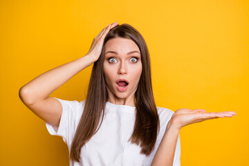 Photo portrait of shocked girl with open mouth holding blank space in hand touching head isolated on bright yellow colored background