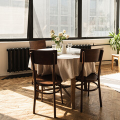 Bright living dinning room interior design. Windows, round dinner table, chairs, flowers bouquet on linen tablecloth. Warm sunlight shadows on the wooden parquet. Retro vintage bohemian home concept.