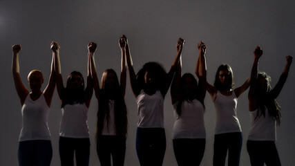 Silhouetted group of diverse women holding, raising their arms together while standing over grey...
