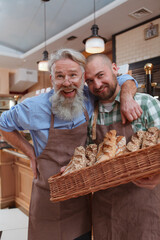 Vertical shot of a cheerful senior male baker and his son laughing posing with basket of freshly baked bread