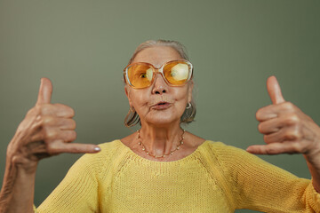 Very emotional elderly lady showing shaka sign and looking at camera. Skinny pop eyed old woman in casual yellow shirt posing isolated over green blackground. Orange glasses. Gray short hair.