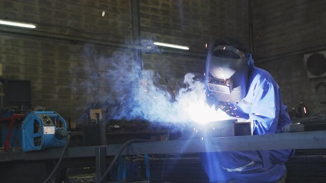 Asian male technician worker in a dressed in protective uniform welding metal frame wear welders leathers, electric welding on steel structure in factory workshop