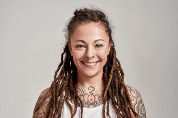 Close up portrait of young tattooed caucasian woman with dreadlocks wearing white shirt, smiling at...