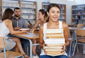 Smiling female student sitting with pile of books in high school library on background with working coursemates