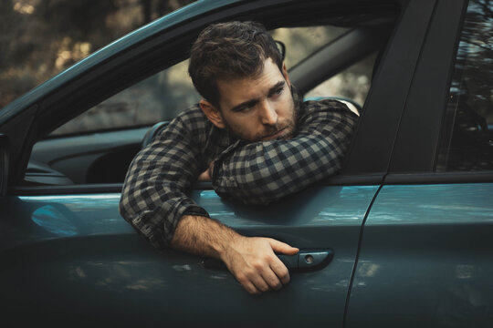 Young man with light skin and light eyes posing leaning out of the car window sticking his arms out and looking back, car parked on forest road, wears plaid shirt.