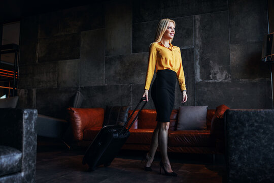 Full Length Of Middle-aged Blonde In Formal Wear Pulling Suitcase And Leaving The Hotel.