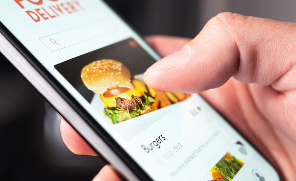 Food Delivery App Order With Phone. Online Mobile Service For Take Away Burger And Pizza. Hungry Man Reading Restaurant Menu, Website And Reviews With Smartphone. Takeout Or Fast Courier Deliver.