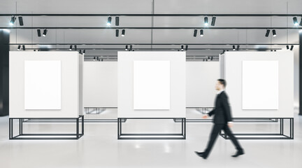 Businessman walking in exhibition room with three blank banners on gray wall.