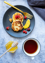Cottage cheese pancakes, Russian syrniki, on a black plate with berries, orange and cup of tea. A jar of honey and honey stick. Healthy breakfast rich in calcium.