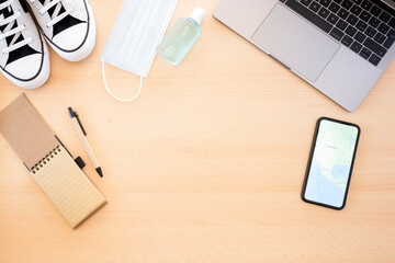 Flat lay, travel concept during Covid-19 with copy space. A face mask, hand sanitizer, shoes, notepad, laptop and a smart phone showing the United States map are arranged on a wooden table. 