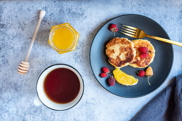 Cottage cheese pancakes, Russian syrniki, on a black plate with berries, orange and cup of tea. A jar of honey and honey stick. Healthy breakfast rich in calcium.