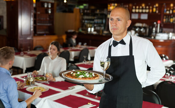 Polite waiter holding tray in restaurant with customers behind him..