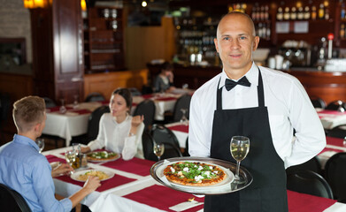 Fototapeta na wymiar Polite waiter holding tray in restaurant with customers behind him..