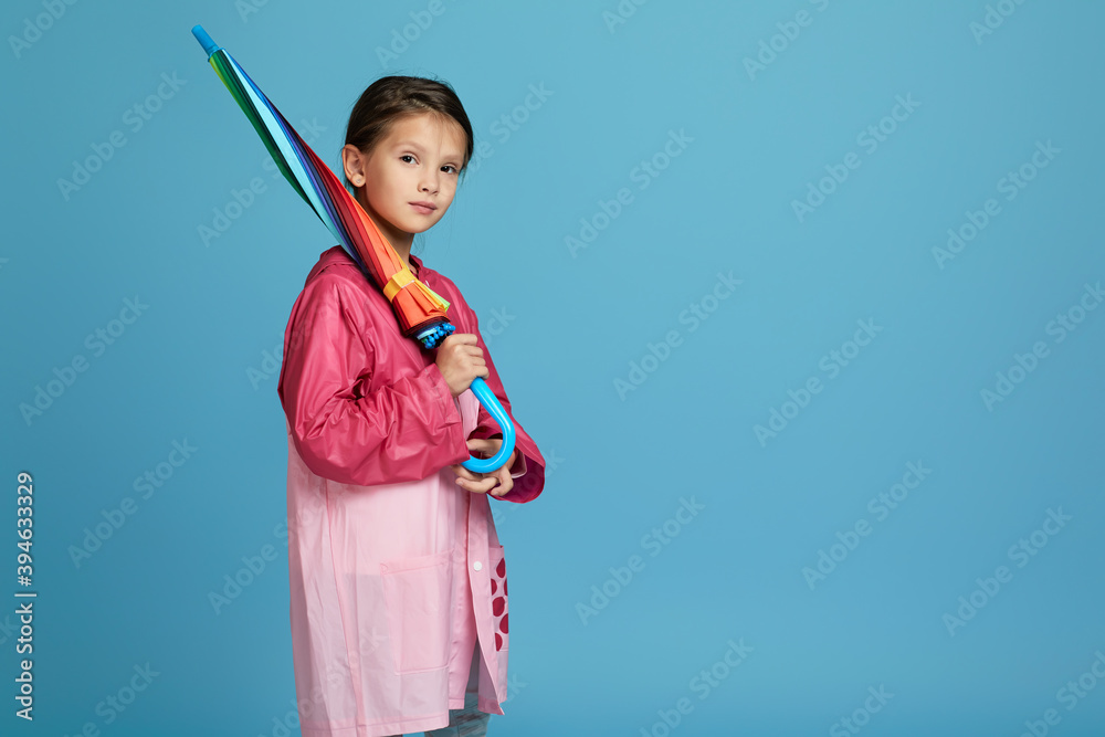 Wall mural cute child girl with multicolored umbrella in pink rain coat on blue background. Copy space for text