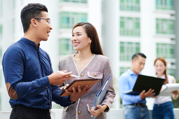 Positive young Vietnamese business people standing outdoors and discussing articles on tablet computer