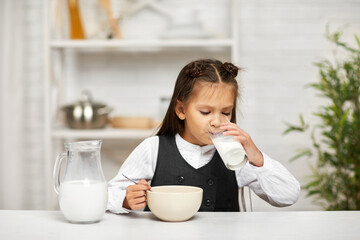 smiling cute little girl in school uniform having breakfast: cereal with the milk. child drinking...