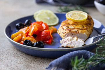 Oven baked or grilled chicken breast with baked bell pepper, lemon and olives, light concrete background. Healthy food.