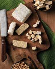 Top View Cut Slicing Raw Tempeh on Wooden Chopping Board, Above Rustic Brown Table. With Banana Leaf, Knife, and Soy Bean