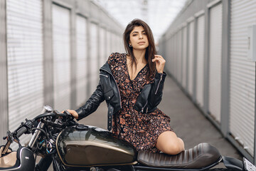 Fototapeta na wymiar Portrait of a bright and lovely female model in a leather jacket and dress posing next to a black motorcycle and looking straight into the camera against the backdrop of white walls. Brutality concept