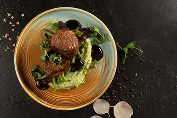 Juicy fried fillet mignon steak with asparagus bean puree and berry sauce. A plate with a hot meat dish, spices and herbs on a black stone kitchen table.