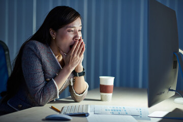 Pretty tired young Vietnamese businesswoman yawning when reading e-mails on computer screen in dark office