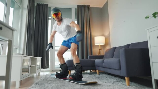 Fun video. Man in shorts and a T-shirt depicts snowboarding on a carpet in a cozy room. Waiting for a snowy winter