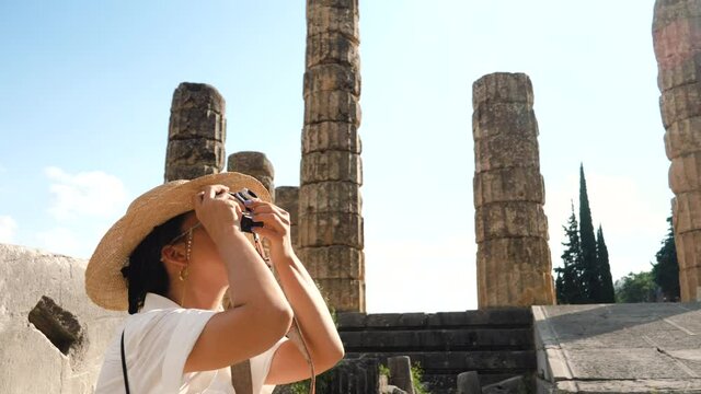 Young woman taking pictures at ruins of temple of Apollo in Delphi, archaeological site by mount Parnassus, Greece, Europe. Vintage camera, large hat, fashion white dress.
