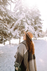 Beautiful woman in hat, plaid scarf and coat posing with joy outside in the snow forest. Cheerful curly lady enjoying winter moments in a snowy park. Holidays, season and leisure concept. Christmas.