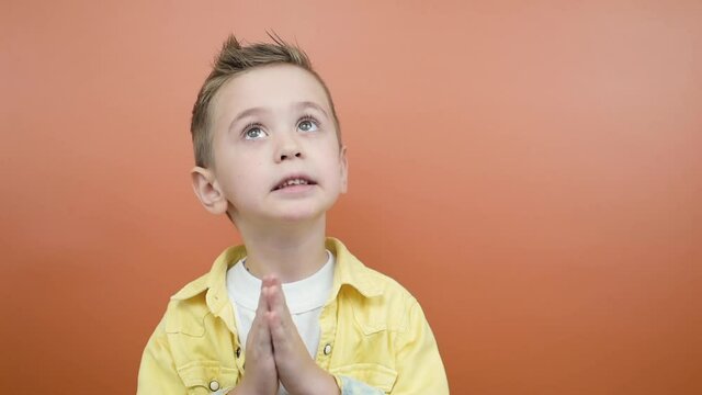 Adorable kid in a yellow shirt is asking for something while doing pray gesture and say please standing isolated over a orange background