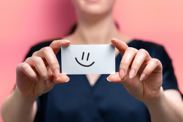 A blurry woman holds a white card with a smiling emoticon drawn on it. Close up. Pink background