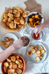 Delicious breakfast with tiny mini pancakes, cherries, apricots, jam on a white table. Small children hold pancakes in their hands. view from above