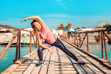An adult happy woman in sports clothes is engaged in stretching. Pier and beach in the background. Sports and healthy lifestyle