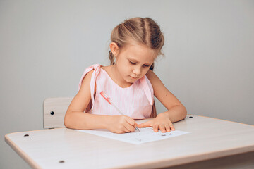 Portrait of a concentrated adorable child in a dress sitting at a white table and writing numbers in a special sheet of paper. Study concept. photo with noise