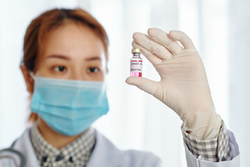 Young serious Vietnamese female doctor in medical mask looking at vial with vaccine against coronavirus