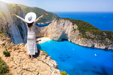 A happy tourist woman in a white summer dress and with hat enjoys the spectacular view to the famous shipwreck beach, Navagio, on the island of Zakynthos, Greece