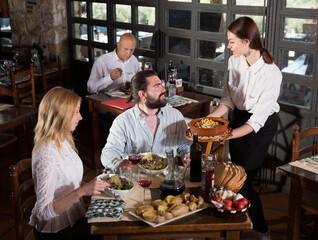 Fototapeta na wymiar Young attractive waitress bringing delicious meals to visitors of cosy rustic style restaurant