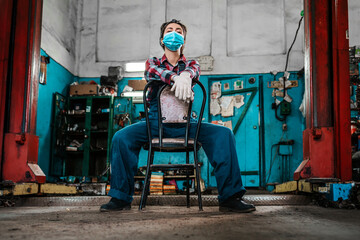 A young female mechanic in uniform, wearing gloves and a medical mask, poses sitting on a chair.In the background there is an auto repair shop. Bottom view