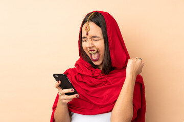 Young Indian woman isolated on beige background with phone in victory position