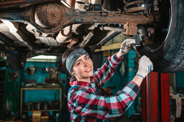 Fototapeta na wymiar Portrait of a young beautiful smiling female mechanic in uniform and gloves who repairs a car. The car is on the lift